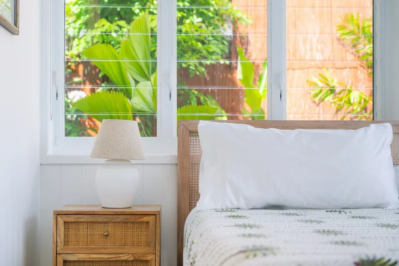 Bright bedroom detail showing rattan bedside table, ceramic lamp, and lush tropical garden visible through louvre windows at Havana, Port Douglas