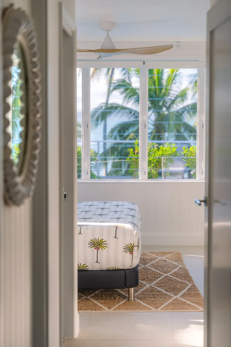 View through a doorway into a sunlit bedroom at Havana Port Douglas with louvre windows framing swaying palm trees, circular decorative mirror on the wall