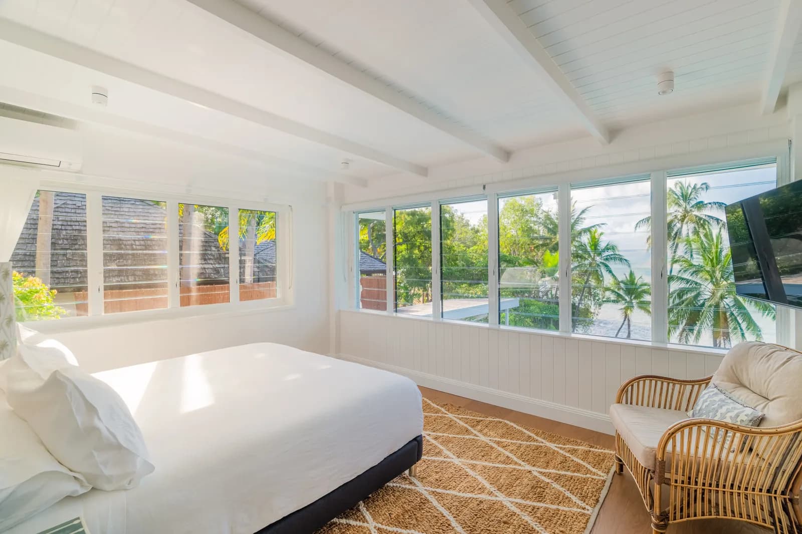 Bright master bedroom at Havana Port Douglas with king bed, white linens, exposed beam ceiling, rattan armchair, jute rug, and wrap-around windows with palm tree and pool views