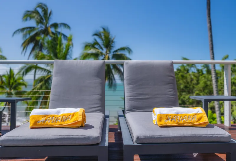 Close-up of two pool loungers at Havana Port Douglas with yellow branded Havana towels, palm trees, and blue sky in the background