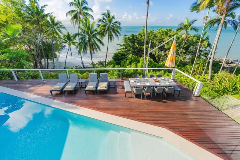 Wide view from Havana Port Douglas pool deck showing loungers, outdoor dining setting with orange umbrella, and a sweeping panorama of Four Mile Beach and the Coral Sea through coconut palms