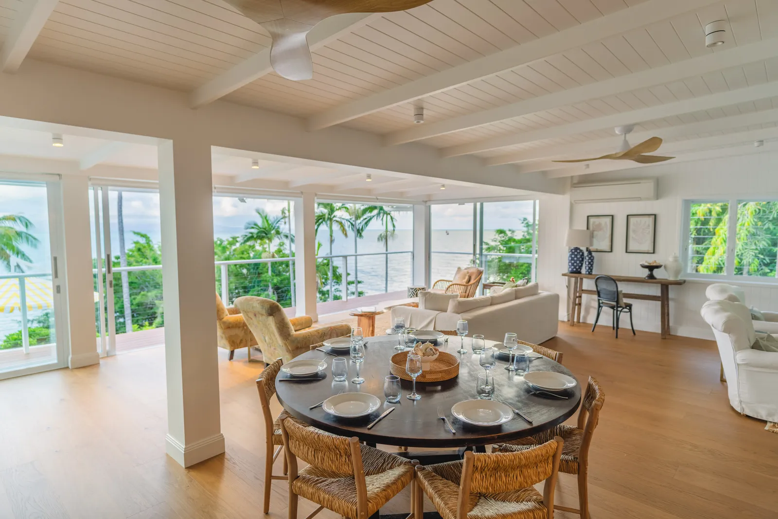 Open-plan dining and living area at Havana Port Douglas with a round black dining table set for six, woven chairs, white sofas, exposed timber ceiling beams, and panoramic ocean views through floor-to-ceiling windows