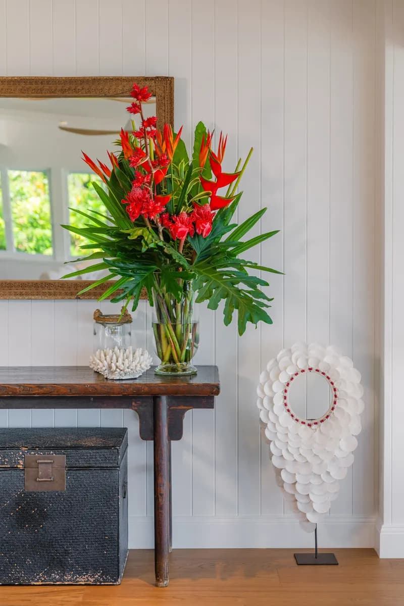 Entry console table at Havana Port Douglas featuring a vibrant red tropical flower arrangement in a glass vase, vintage trunk, coral ornament, and white shell sculpture against white VJ panelling