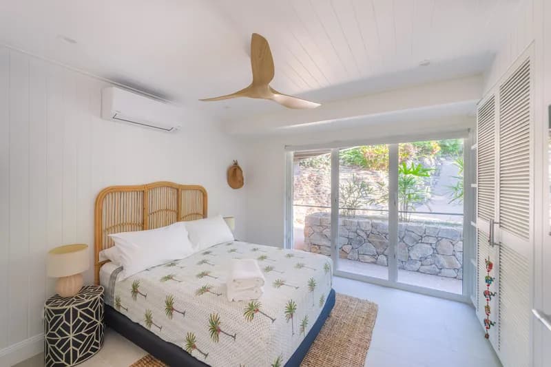 Ground-floor bedroom at Havana Port Douglas with rattan bedhead, palm-print bedspread, timber ceiling fan, jute rug, and sliding doors opening to a stone-walled garden