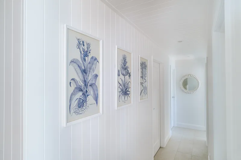 White VJ-panelled hallway at Havana Port Douglas lined with three framed blue-and-white botanical illustrations and a decorative circular mirror
