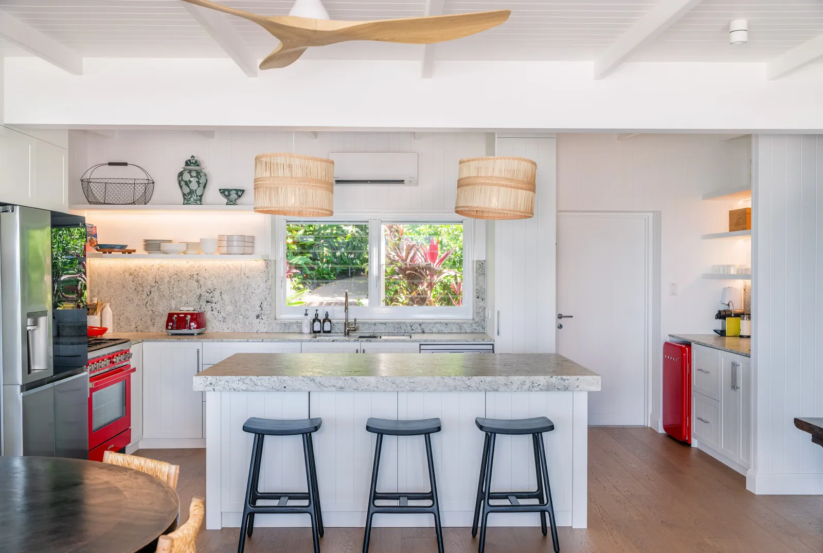 Spacious kitchen at Havana Port Douglas with large stone island bench, black bar stools, rattan pendant lights, red Smeg oven, and garden window