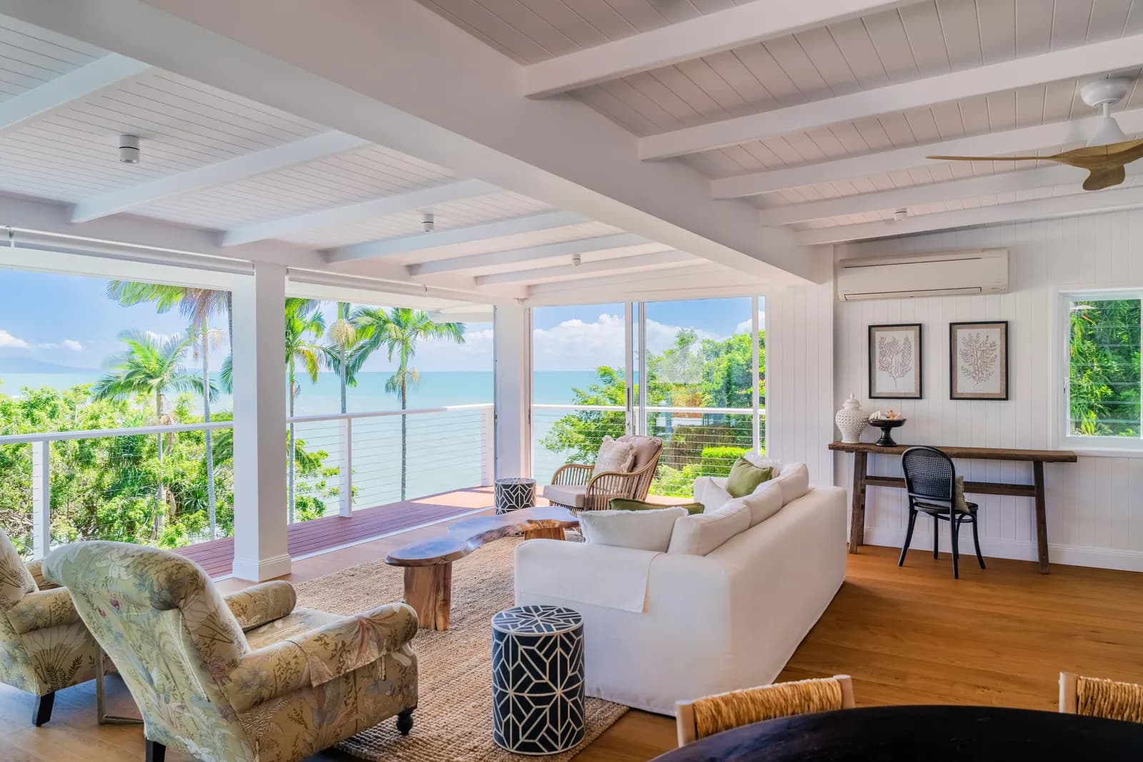 Open-plan living room at Havana Port Douglas with white sofa, rattan armchairs, timber floors, exposed beam ceiling, and floor-to-ceiling windows framing Coral Sea views and palm trees