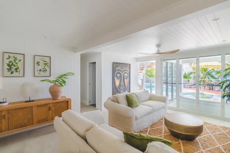 Open-plan living room at Havana Port Douglas with white linen sofas, olive cushions, jute rug, timber sideboard, botanical prints, and glass doors opening to the pool deck