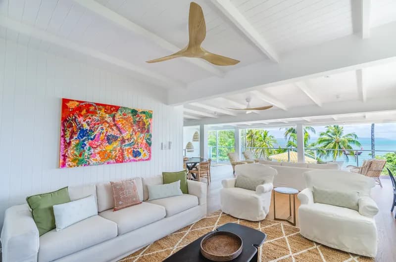 Bright lounge at Havana Port Douglas featuring white slipcovered sofas, a vibrant abstract painting, sculptural timber ceiling fan, and ocean views through glass walls