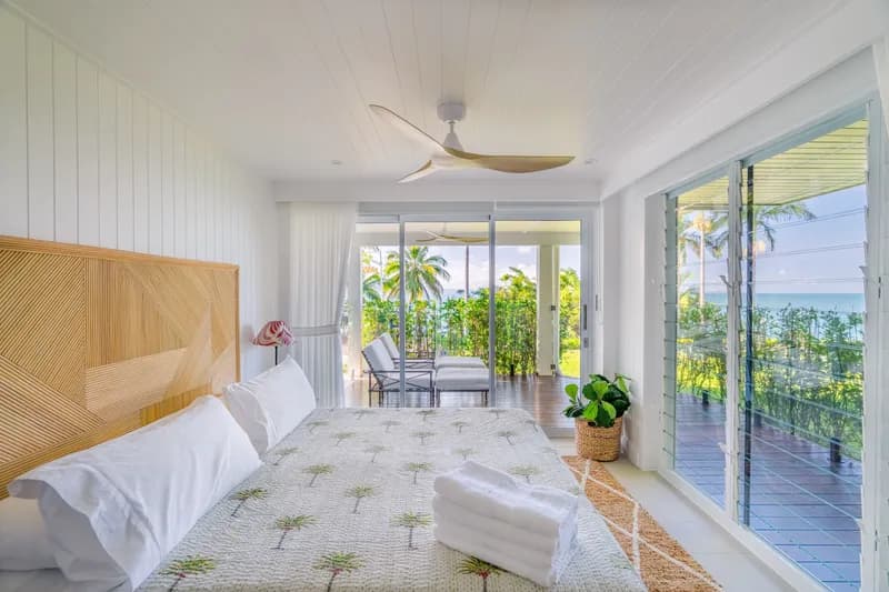 Master bedroom at Havana Port Douglas with geometric rattan bedhead, palm-print bedspread, and floor-to-ceiling glass doors opening to a balcony with ocean and tropical garden views