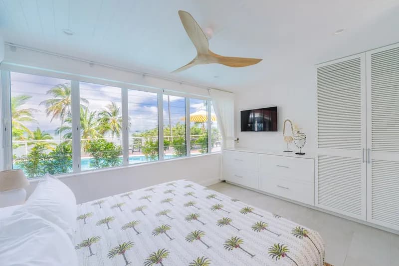 Light-filled master bedroom with palm-print bedding, ceiling fan, wall-mounted TV, and panoramic views of the pool and palm trees at Havana, Port Douglas