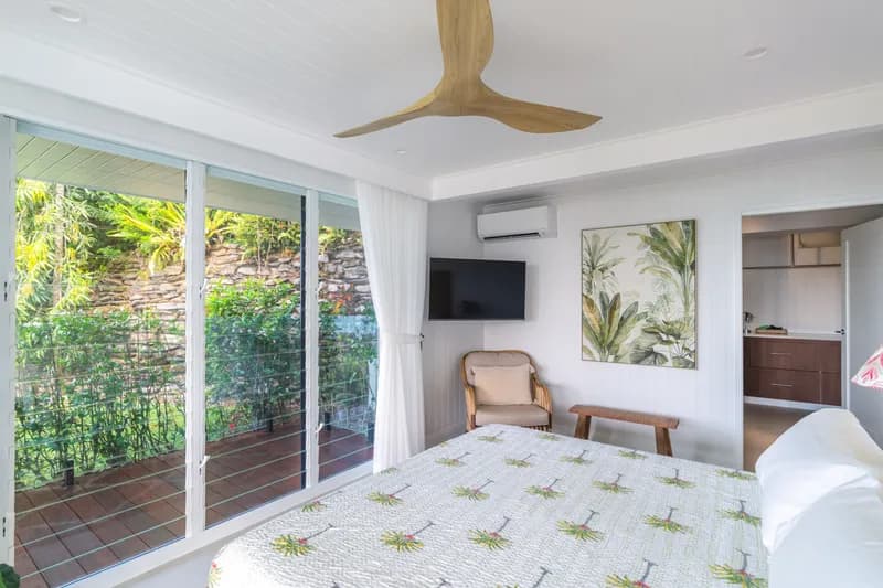 Master bedroom at Havana Port Douglas showing wall-mounted TV, tropical botanical artwork, rattan armchair, sliding glass doors to a private balcony with lush garden views, and ensuite entry