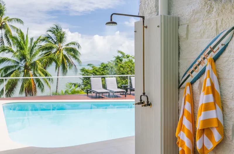 Outdoor brass shower beside the pool with striped towels, a surfboard, and palm trees overlooking the Coral Sea at Havana, Port Douglas