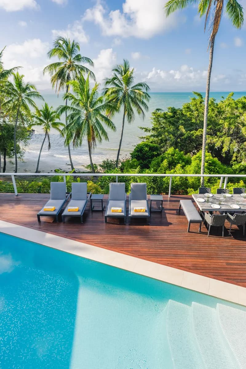 Pool and timber deck at Havana Port Douglas with sun loungers and outdoor dining table overlooking Four Mile Beach, coconut palms, and the turquoise Coral Sea