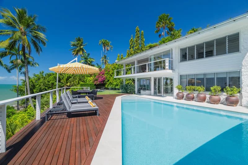 Swimming pool and timber sun deck at Havana Port Douglas with loungers, striped yellow umbrella, terracotta pots, and Coral Sea views framed by palm trees