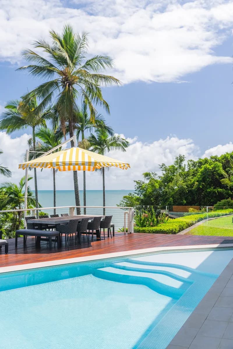 Sparkling pool and timber deck with outdoor dining table under a yellow-striped umbrella, palm trees framing Coral Sea views at Havana, Port Douglas