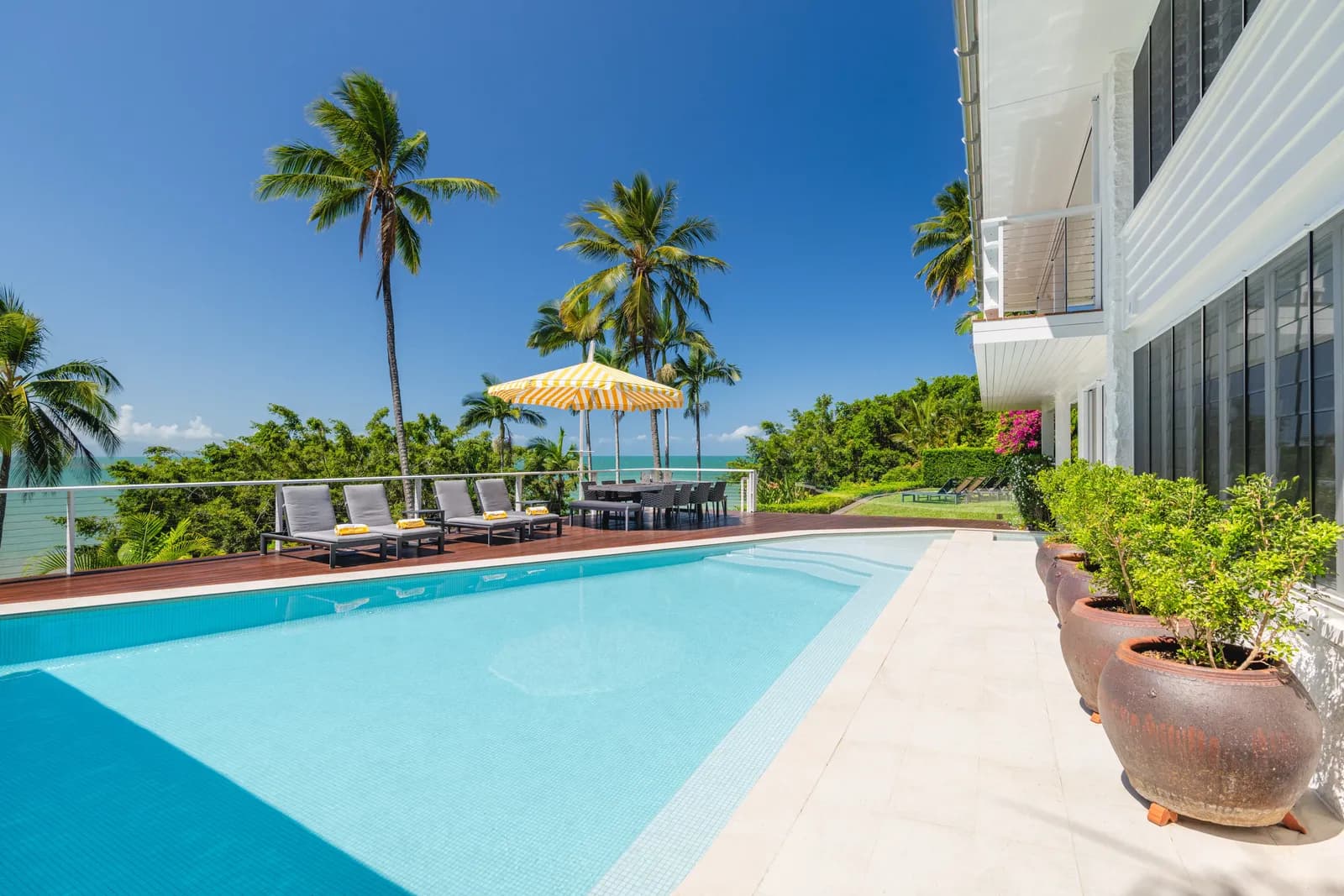 Sparkling pool at Havana Port Douglas with outdoor dining setting, sun loungers under a yellow striped umbrella, coconut palms, and turquoise ocean beyond
