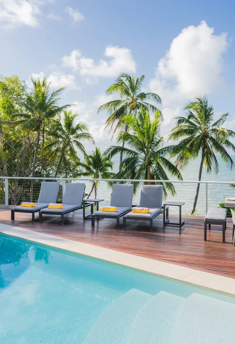 Four sun loungers with yellow towels on the timber pool deck at Havana, Port Douglas, with palm trees and ocean beyond
