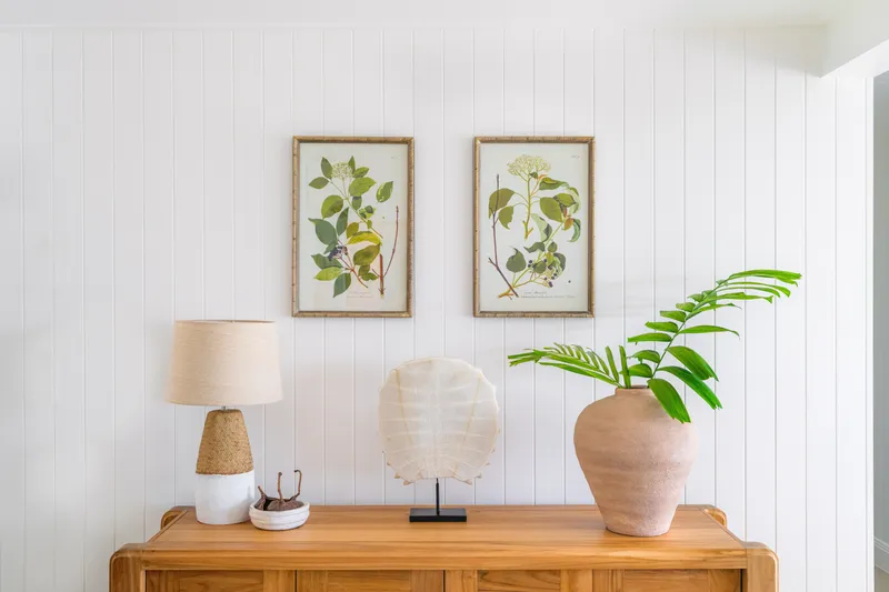 Timber sideboard styled with a woven lamp, shell disc sculpture, and terracotta vase with palm frond beneath framed botanical prints on white VJ walls at Havana Port Douglas