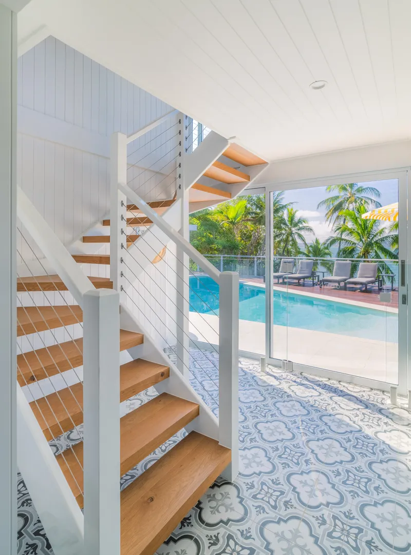 Timber and wire-balustrade staircase at Havana Port Douglas with decorative blue-and-white patterned floor tiles and glass doors framing the pool, palm trees, and ocean beyond