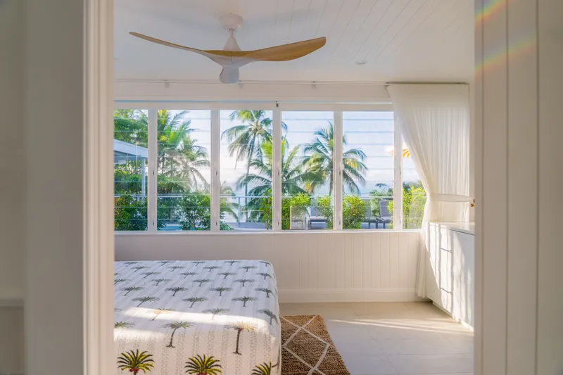 Bright upper-level bedroom at Havana Port Douglas with wide louvre windows overlooking coconut palms, white timber ceiling, and natural light streaming across the palm-print bed
