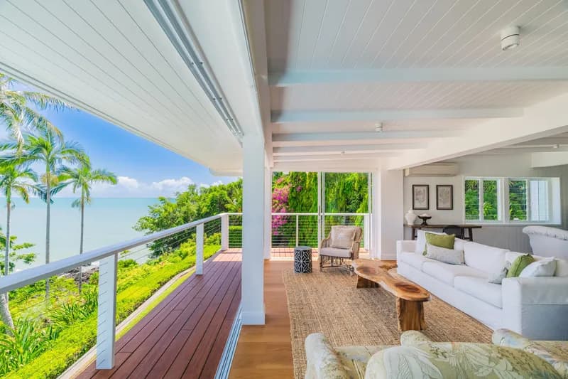 Covered verandah and open-plan living area at Havana Port Douglas with timber deck, white sofa, jute rug, and sweeping views over tropical gardens to the Coral Sea